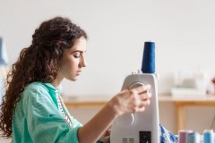 Young seamstress with dark curly hair in colorful shirt dreamily using sewing machine in sewing workshop